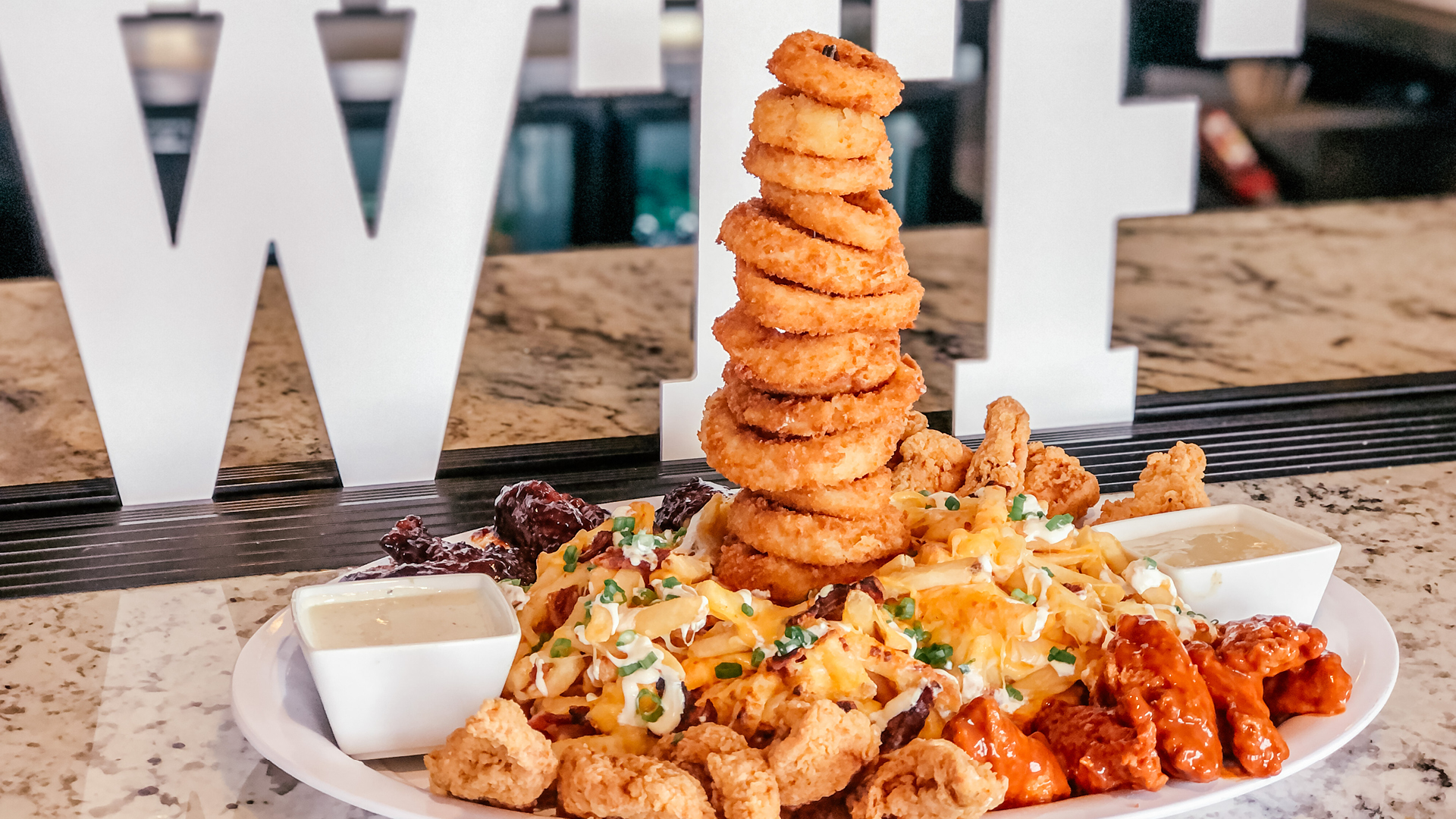 An image of stacked fried onion rings on an appetizer dish at PINSTACK with wings, fries, and dipping sauces