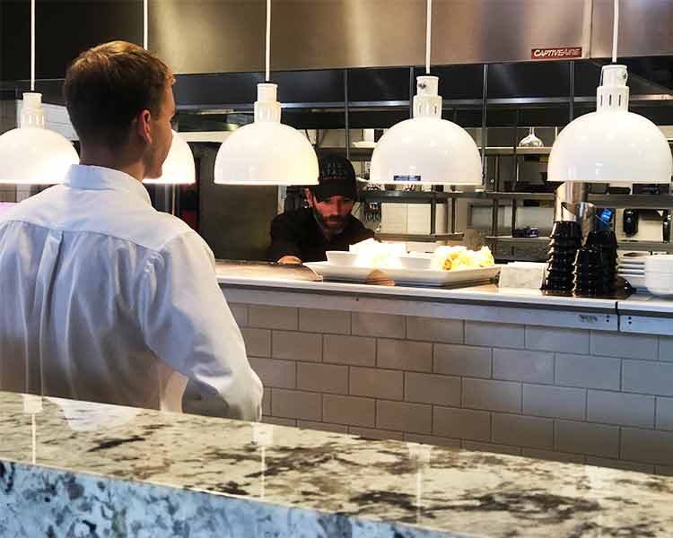 Image looking into restaurant expo with line cooks preparing dish as server looks on. 