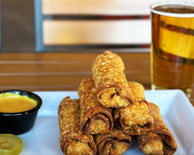 A photo of the new delicious cheeseburger egg rolls served with a side of special sauce on our climate-controlled patio table with a glass of beer.