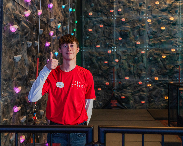 Photo of a Team Member in a red shirt giving the thumbs up sign in front of rock wall.