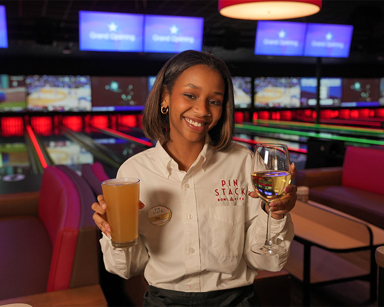 Photo of a server smiling and holding two beverages she is delivering to a lane-side guest. 