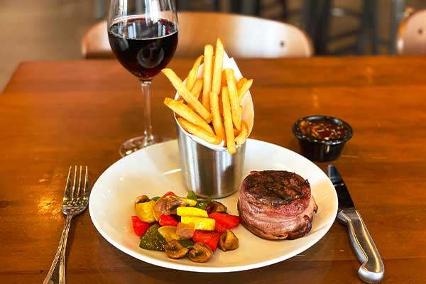 Filet mignon with fries, roasted vegetables, and a glass of red wine served on a wooden table at PINSTACK.
